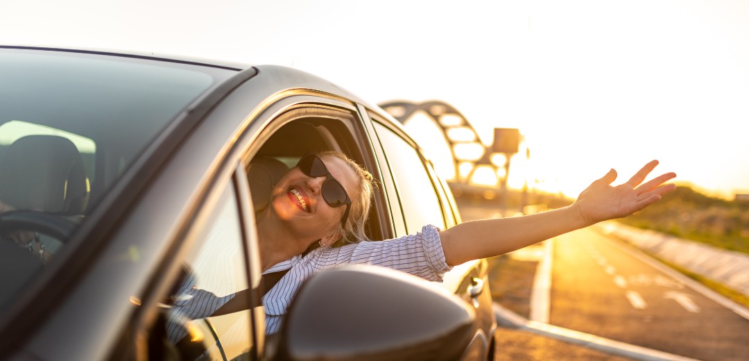 Eine junge blonde Frau fährt in glücklich in der Sonne ihr Auto, trägt eine Sonnenbrille, streckt einen Arm aus dem Fahrerfenster und lacht.