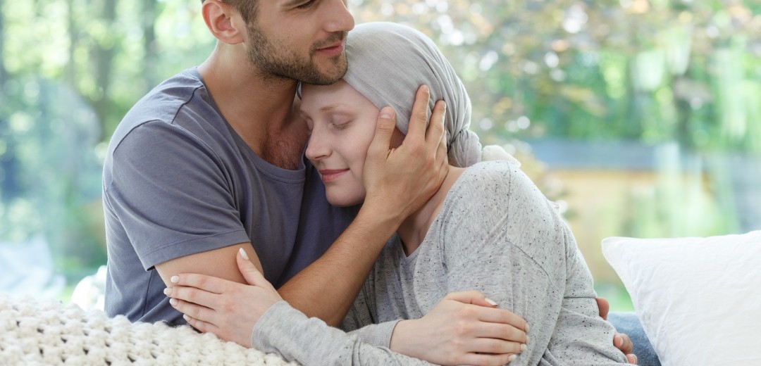 Ein junger Mann hält seine krebskranke Freundin im Arm. Sie sitzen zusammen auf dem Sofa. 