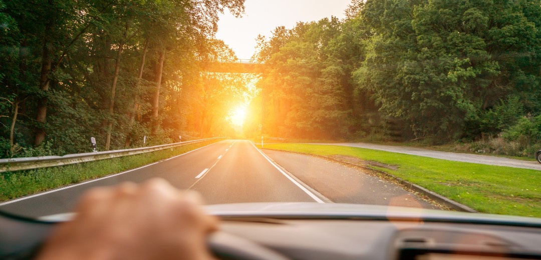 Eine zusehende Hand am Lenkrad eines Autos. Das Auto fährt eine Landstraße im Sonnenuntergang entlang. 