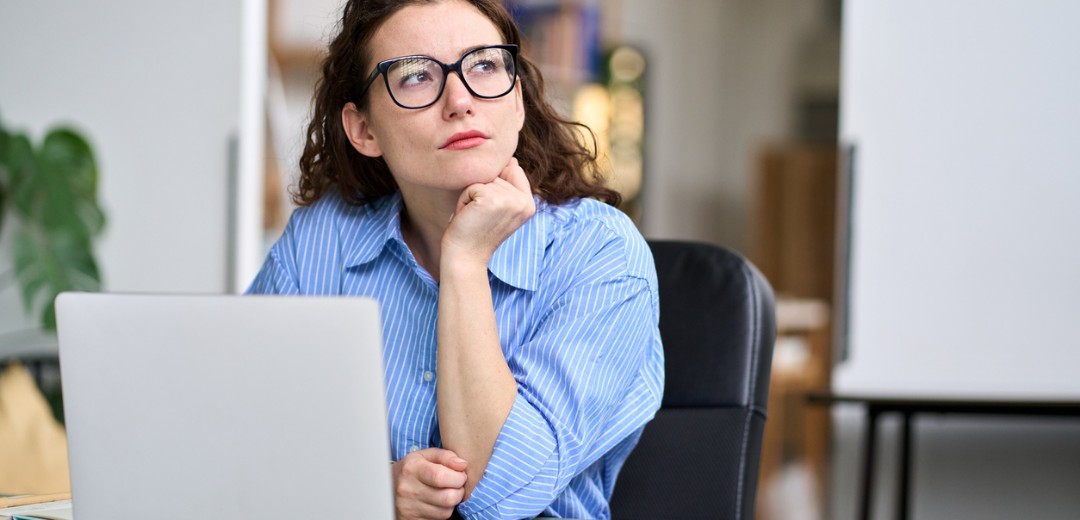 Nachdenkliche Frau mit Brille sitzt am Schreibtisch vor einem Laptop, stützt ihr Kinn auf die Hand und schaut zur Seite.