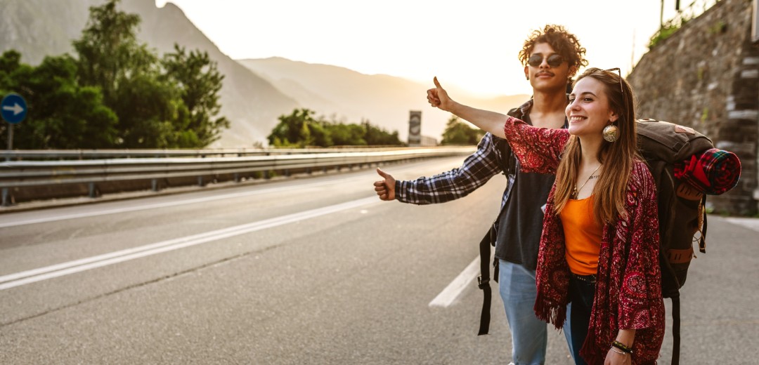 Zwei Backpacker stehen mit ihren Rucksäcken am Straßenrand und halten ihren Daum raus. Sie warten auf eine Mitfahrgelegenheit. 