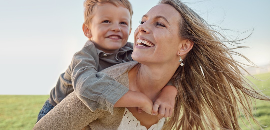 Mutter trägt ein Kind auf dem Rücken in einer sonnigen, grünen Umgebung.