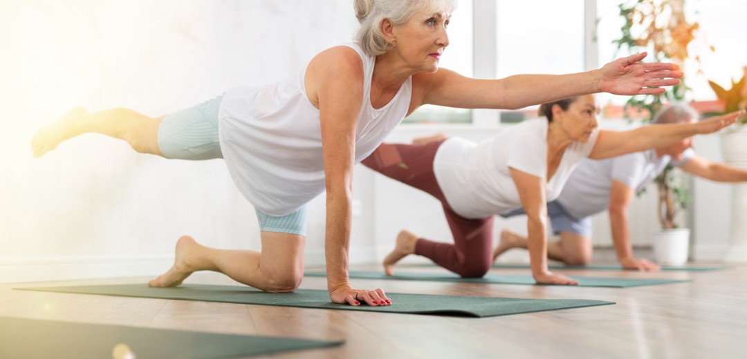 Ältere Frau macht mit anderen eine Balance-Übung auf Yogamatten in einem hellen Raum.