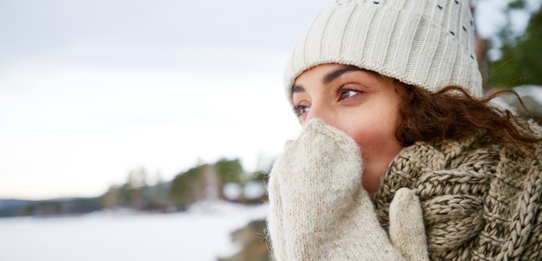 Eine warm eingepackte Frau mit Mütze, Schal und Handschuhen steht draußen in einer verschneiten Landschaft und hält die Hände vor ihr Gesicht, um sich zu wärmen.