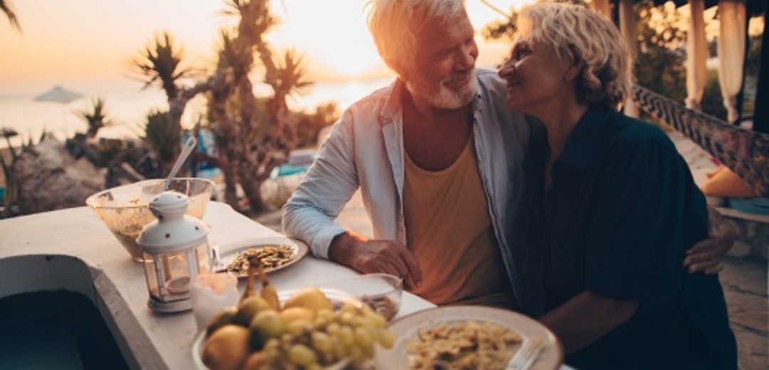Ein älteres Ehepaar (Rentner) genießen ihr Abendessen unter Palmen im Sonnenuntergang. Sie schauen sich läacheldn an.