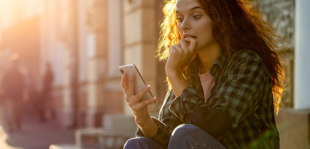 Eine junge Frau sitzt draußen auf einer Steinstufe und schaut gespannt in ihr Smartphone. 