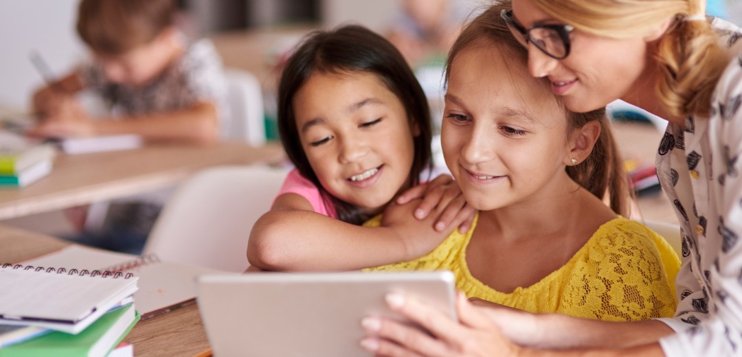 Eine Lehrerin zeigt zwei jungen Schülerinnen etwas auf dem Tablet. Sie sitzen in einem Klassenzimmer.