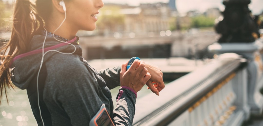 Eine junge Joggerin steht auf einer Brücke und schaut von ihrer Smart Watch hoch. 