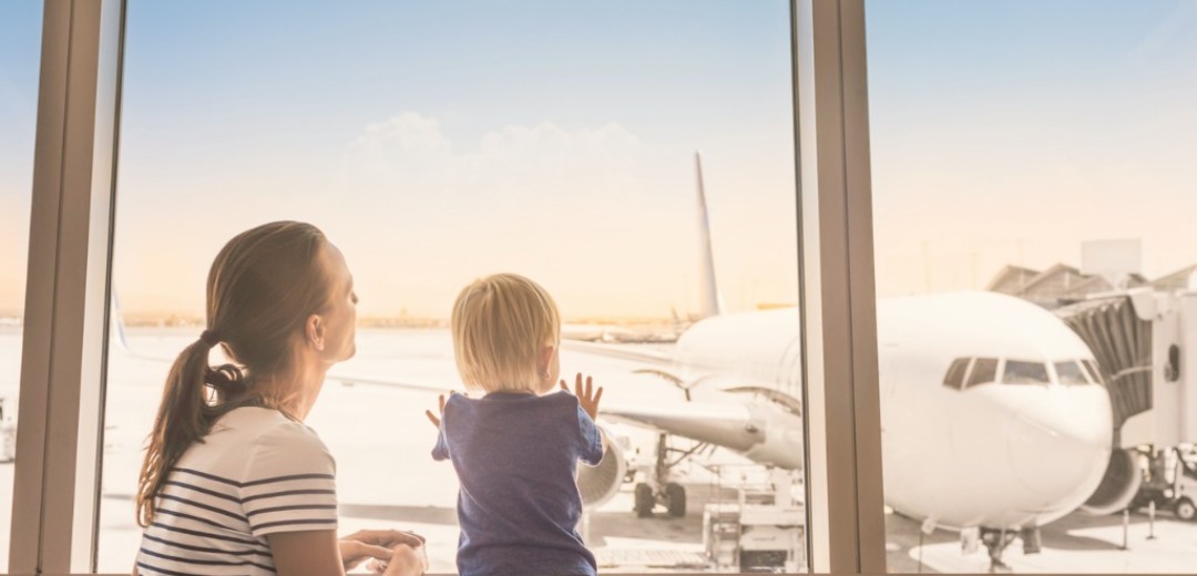 Eine Mutter schaut mit ihrem Kleinkind aus dem Fenster an einem Flughafen. Sie sehen ein Flugzeug, was am Gate steht.