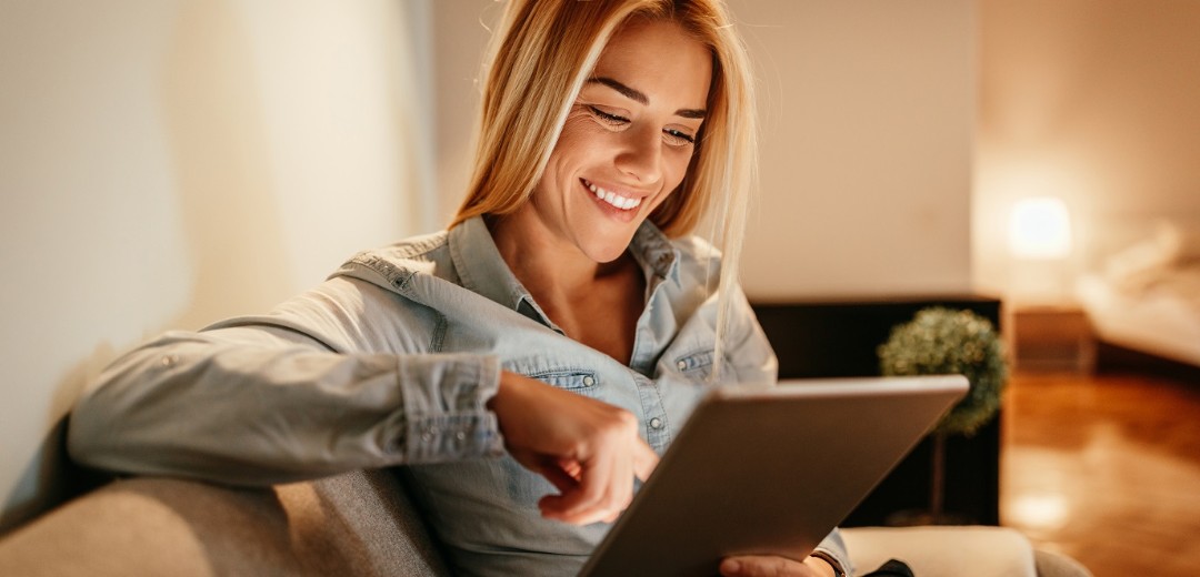 Eine junge Frau lächelt, während sie etwas auf ihr Tablet durchliest. Sie sitzt in ihrem Wohnzimmer auf einer Couch.