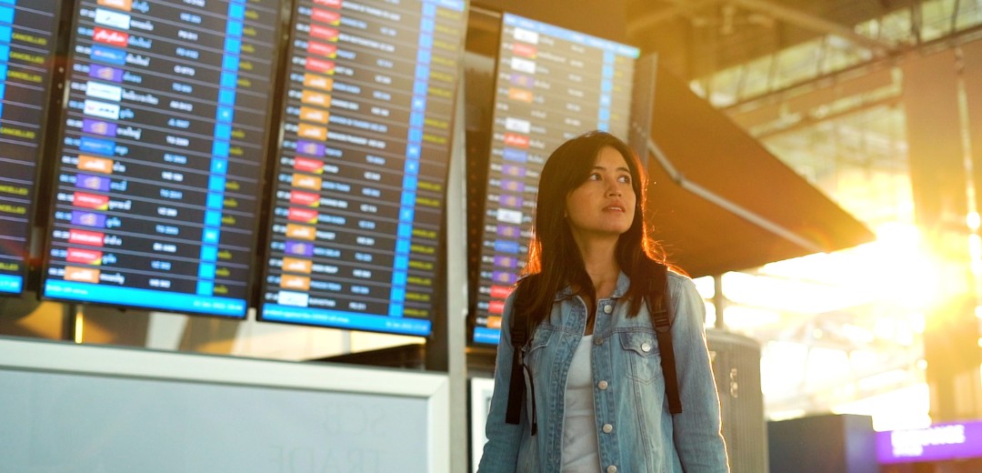 Eine junge Frau steht am Flughafen unter dem großen An- und Abreiseplan und schaut in die Ferne. 