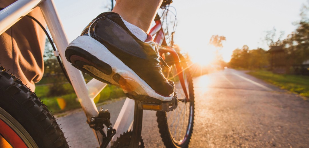 Ein Mann fährt sein Fahrrad im Sonnenuntergang auf einem asphaltierten Weg. Im Fokus stehen seine Sportschuhe.