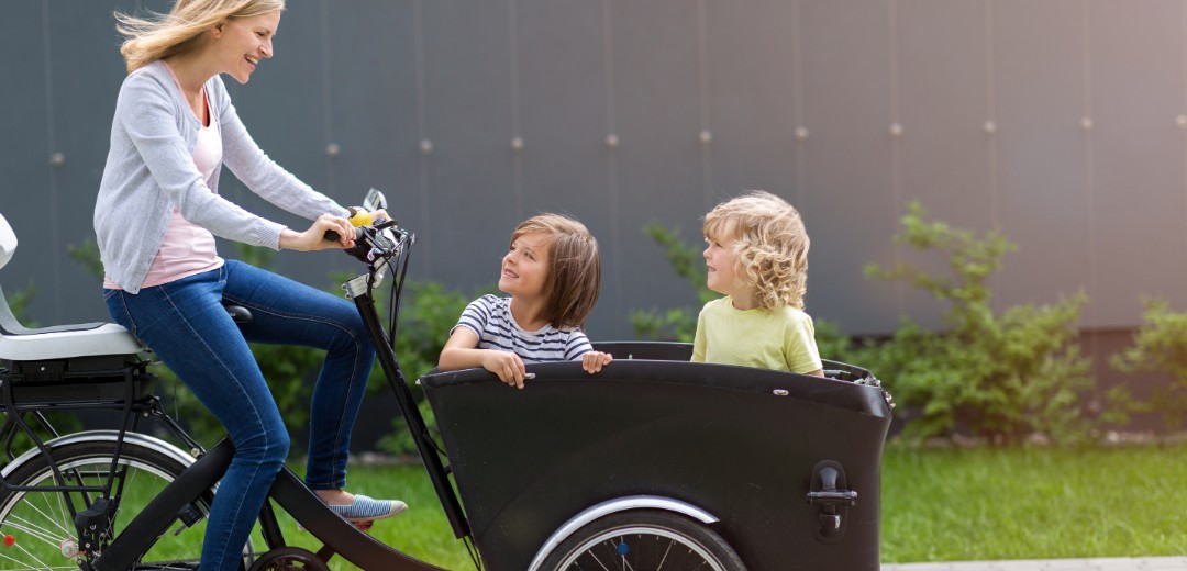 Eine Mutter fährt ein Lastenrad. Vorne im Anhänger sitzen zwei Kinder, die ihre Mutter anschauen. 