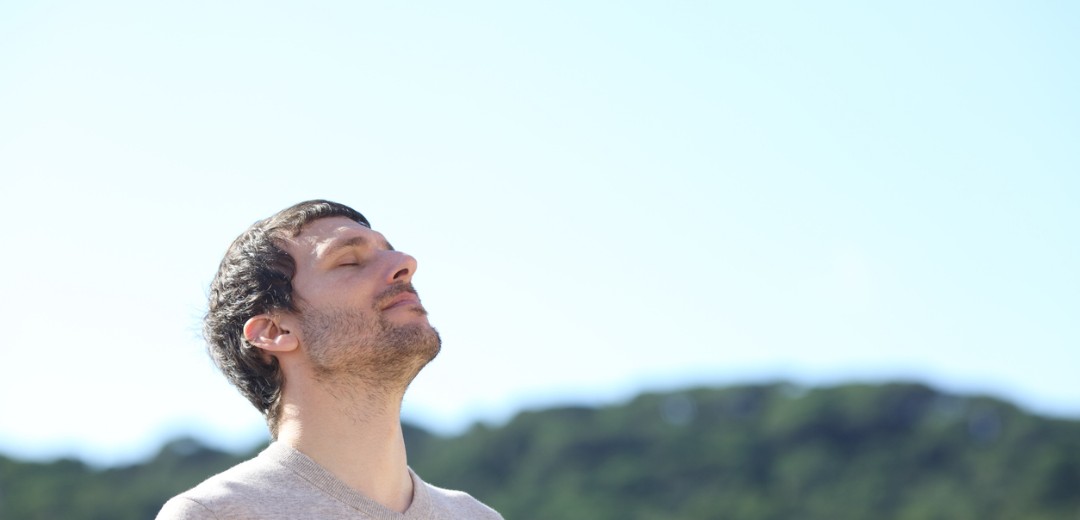 Ein Mann steht draußen mit geschlossenen Augen und hebt das Gesicht leicht in die Sonne, im Hintergrund eine grüne Hügellandschaft.
