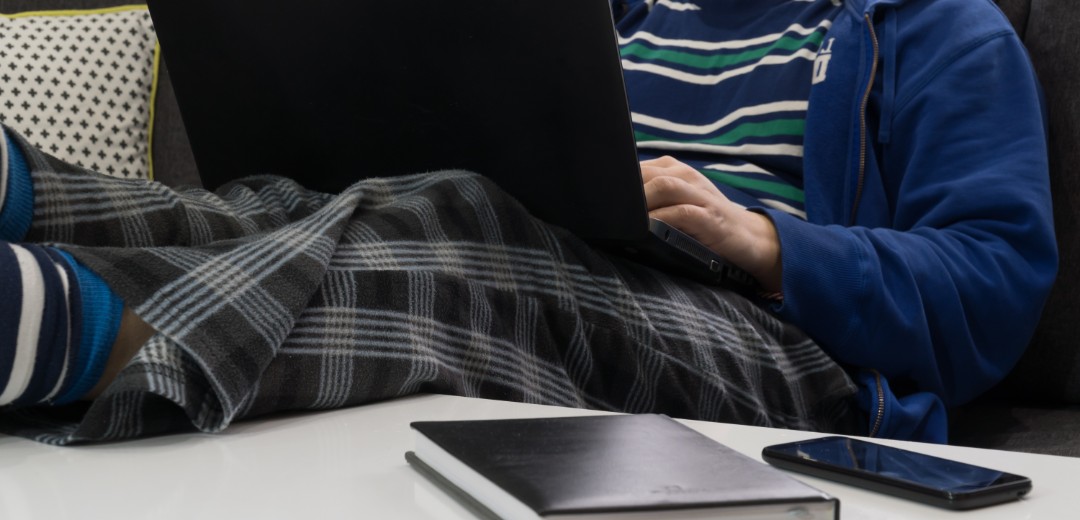 Eine männliche Person (Gesicht nicht zu sehen) liegt auf dem Sofa in Jogginghose und arbeitet am Laptop.