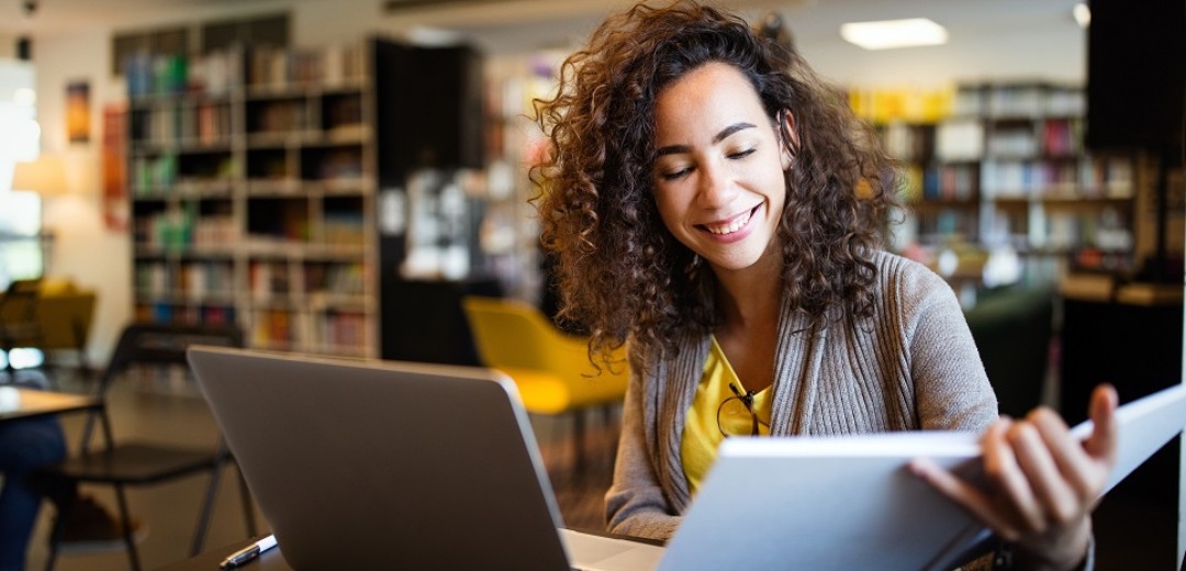 Eine junge Frau sitzt in einem Buchladen am Laptop und schaut währenddessen in ein Buch. 
