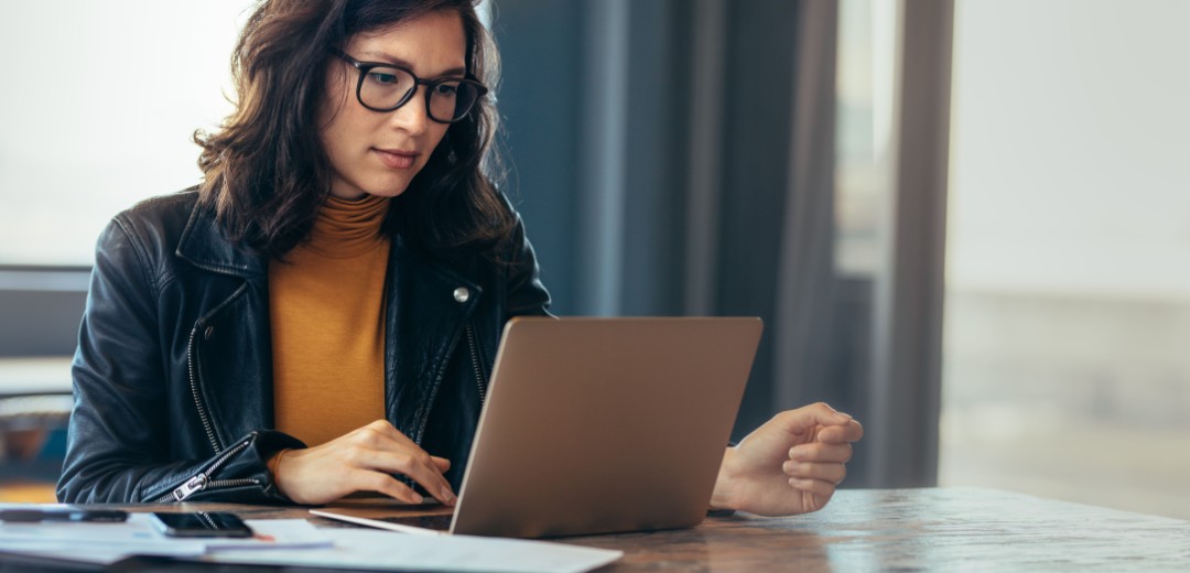 Eine Frau sitzt an einem Tisch und schaut auf ihren Laptop. Neben ihr liegen Dokumente. 