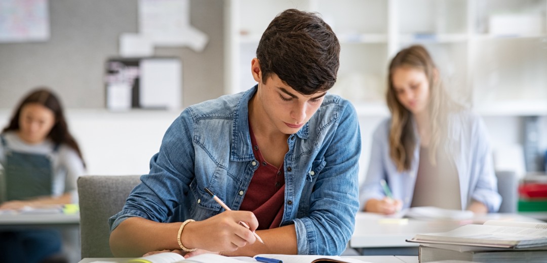 Ein Schüler der Sekundarstufe sitzt im Klassenzimmer an einem Tisch und schreibt etwas in seinen Block