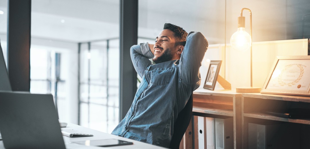 Ein junger Mann sitzt in einem Büro vor dem Computer und lehnt sich mit einem glücklichen Gesichtsausdruck in seinem Stuhl mit den Armen hinter dem Kopf zurück.