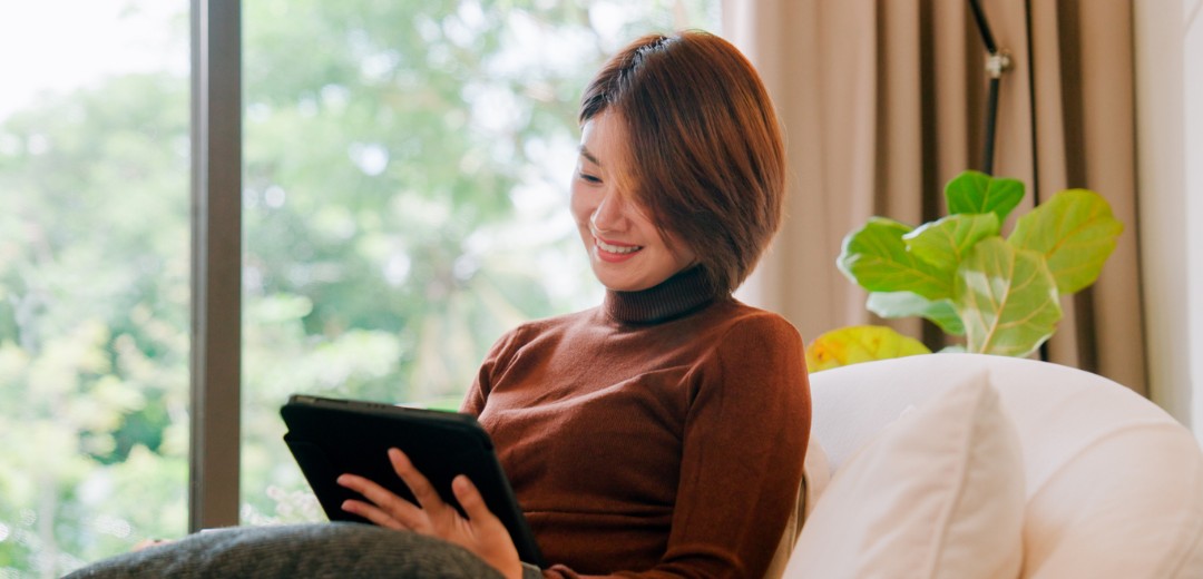 Eine Frau sitzt entspannt auf dem Sofa und schaut auf ihr Tablet