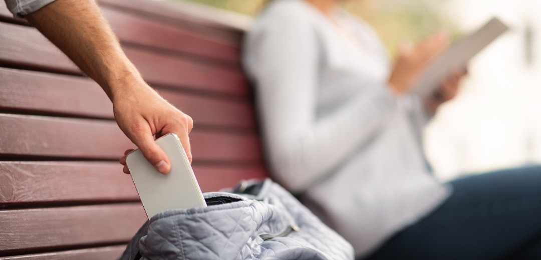 Eine Frau liest ein Buch auf einer Parkbank, während ihr Handy von einem Fremden aus der Tasche gezogen wird. 