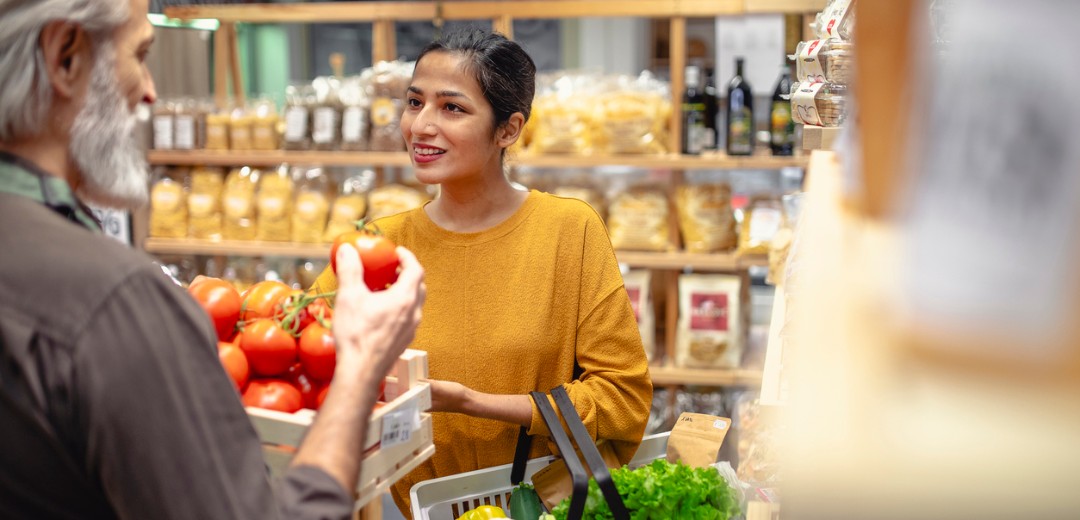 Eine junge Frau lächelt, während sie mit einem älteren Mann in einem Laden spricht und dabei einen Korb mit frischen Lebensmitteln hält. Im Hintergrund sind Regale mit verpackten Produkten zu sehen.