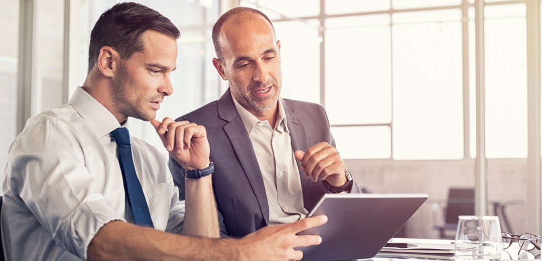 Zwei Business-Männer sitzen zusammen und besprechen etwas am Tablet. 