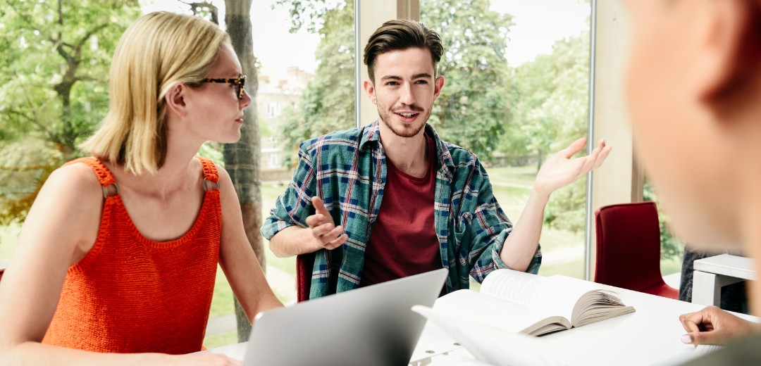 Eine Gruppe von jungen Leuten sitzen zusammen an einem Tisch mit Unterlagen und Laptops. Sie unterhalten sich. 