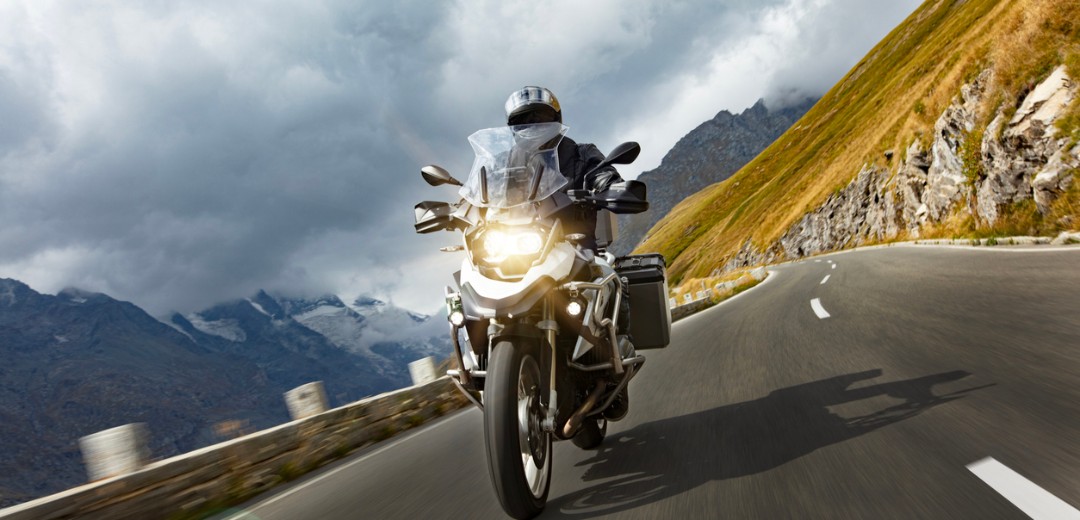 Motorradfahrer fährt auf einer kurvigen Bergstraße, im Hintergrund sind Berge und dunkle Wolken zu sehen.