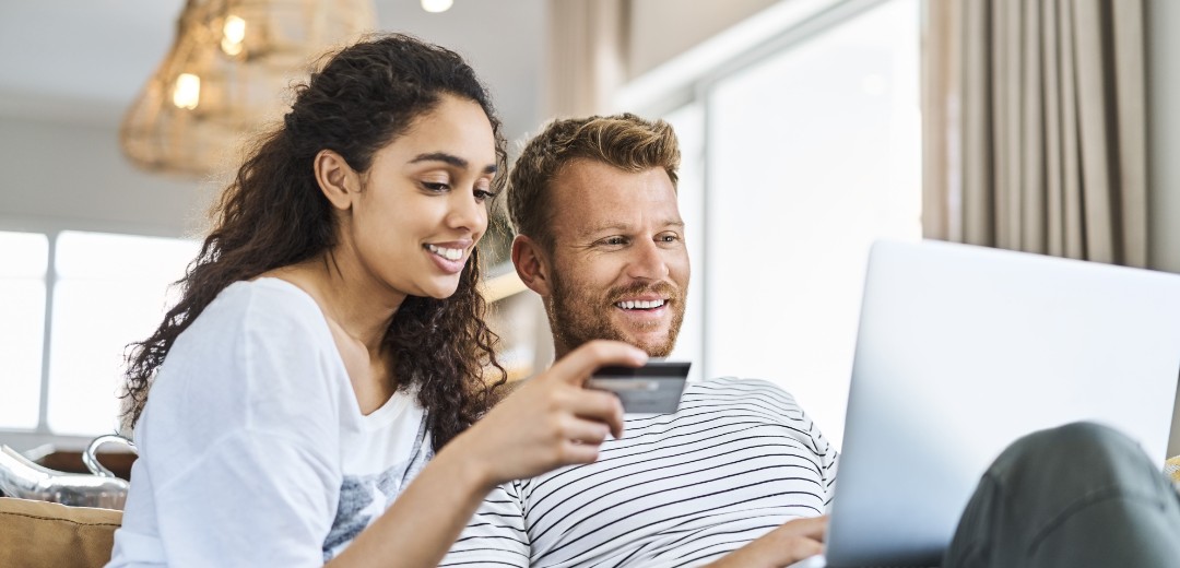 Eine Frau und ein Mann sitzen zusammen auf einem Sofa. Der Mann hält einen Laptop auf seinem Schoß und die Frau hält eine Kreditkarte in der Hand. 