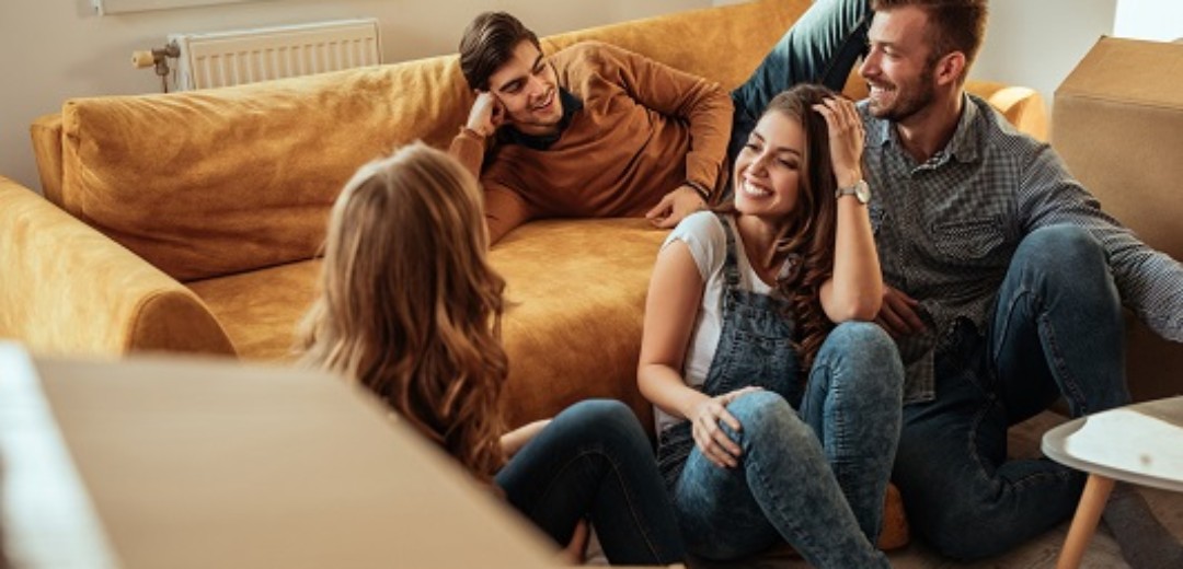 Eine Gruppe von vier Freunden sitzen zusammen zwischen Umzugskartons in einem leeren Wohnzimmer.