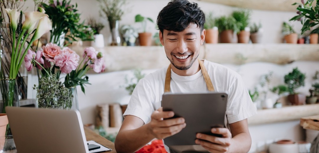 Ein junger Mann sitzt lachend in einem Pflanzenladen am Tablet.