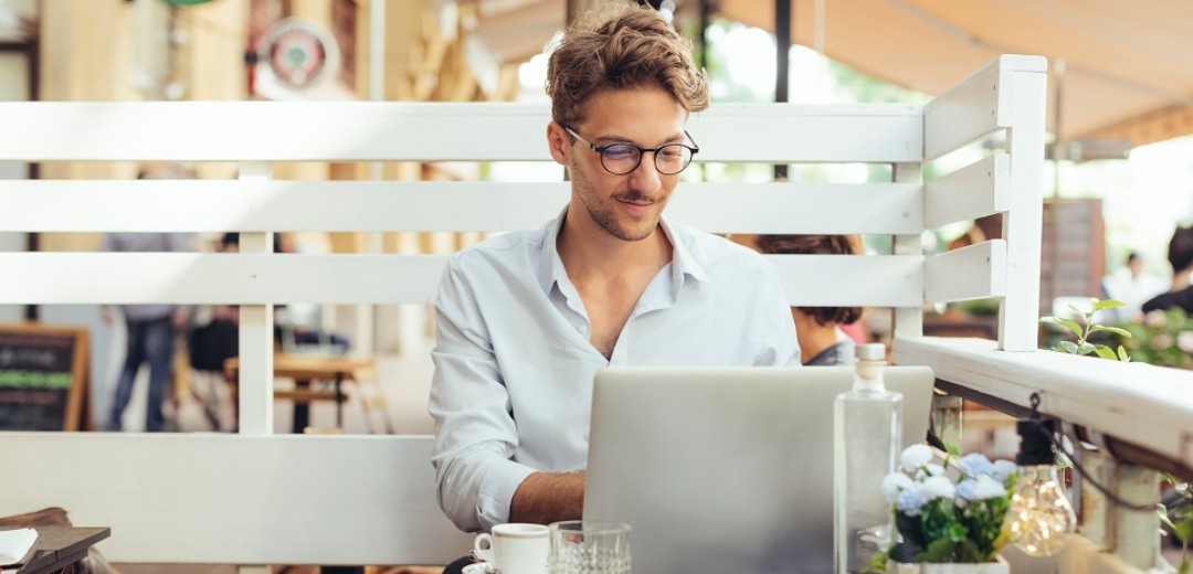 Ein junger Mann sitzt an seinem Laptop und einem Kaffee in einem Café. 