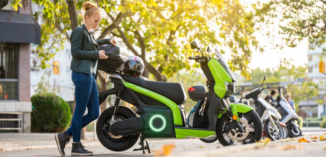 Eine blonde Frau steht neben ihrem elektrischen Motorrad und hält ihren Helm in der Hand. 