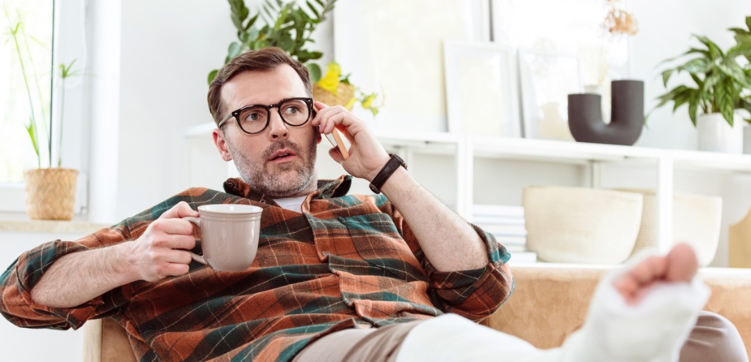 Ein Mann mit Brille sitzt auf einem Sofa, telefoniert und hält eine Tasse. Sein Bein ist in Gips und hochgelegt.
