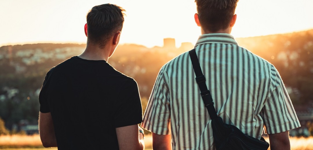Zwei junge Männer stehen im Sonnenuntergang. Sie sind von hinten fotografiert.