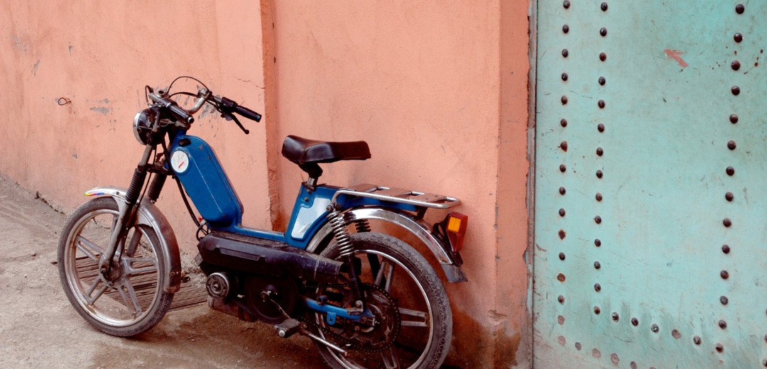 Das Bild zeigt ein älteres Moped, welches an einer Altbau-Hauswand gelehnt ist. 