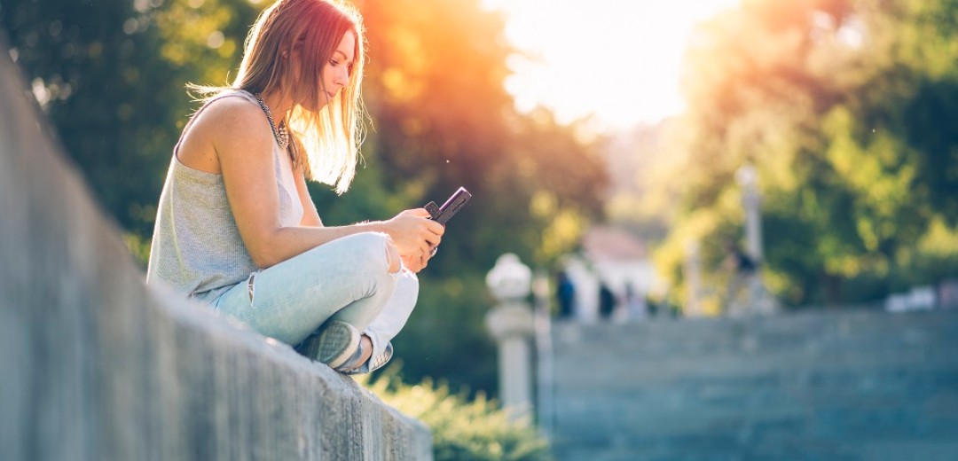 Ein Mädchen sitzt auf einer Steinmauer und schaut auf ihr Mobiltelefon. 