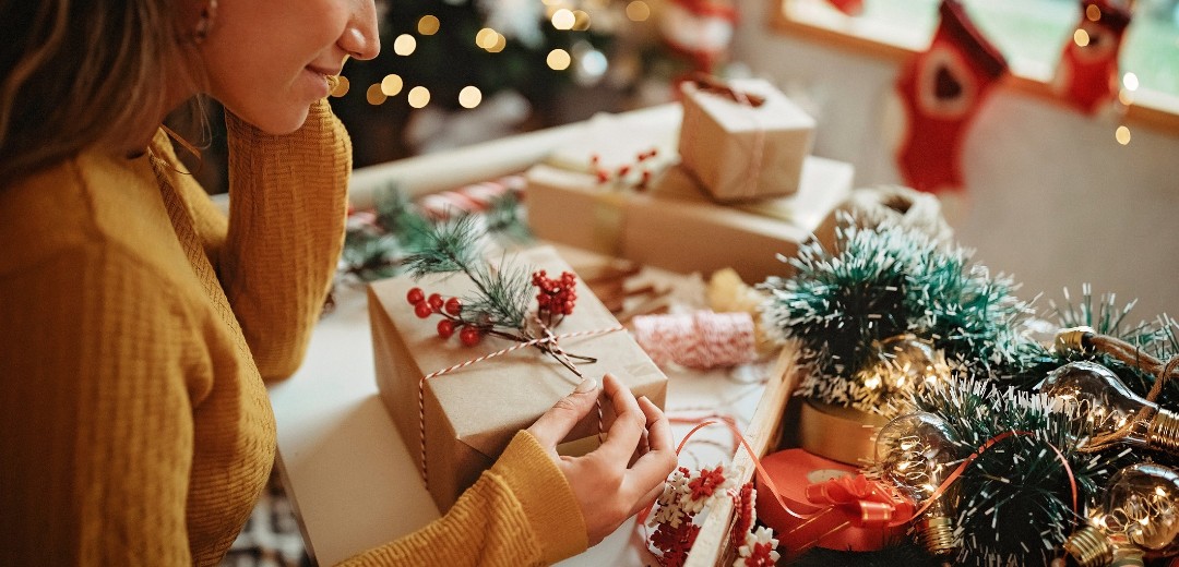Ein Mädchen sitzt nachdenklich am Schreibtisch und begutachtet ein eingepacktes Weihnachtsgeschenk. Um sie herum ist Weihnachtsdekoration und andere eingepackte Weihnachtspäckchen. 