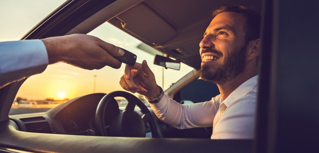 Ein Mann sitzt am Steuer eines Auto und ihm werden die Autoschlüssel gereicht. 