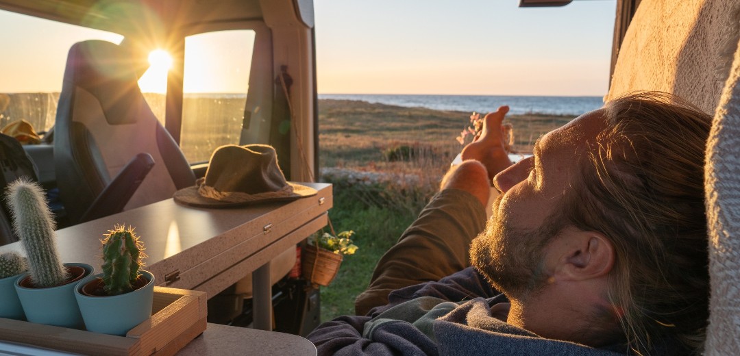 Ein junger Mann liegt in seinem ausgebauten Campervan mit Sonne im Hintergrund.