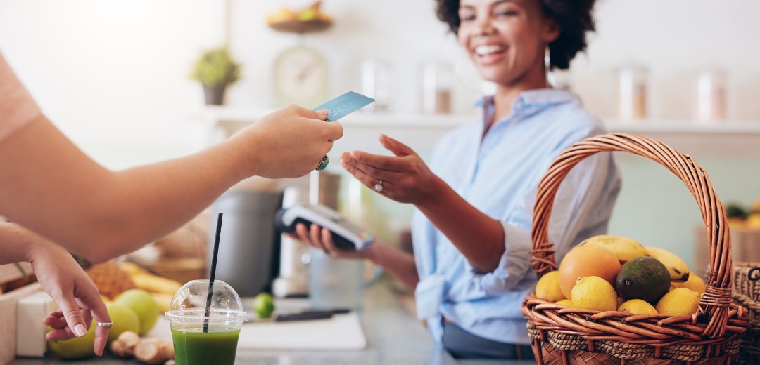 Eine Hand hält einer Frau eine Bankkarte hin. Auf dem Tresen steht ein Smoothie und die Frau hinter dem Tresen kassiert ab. 