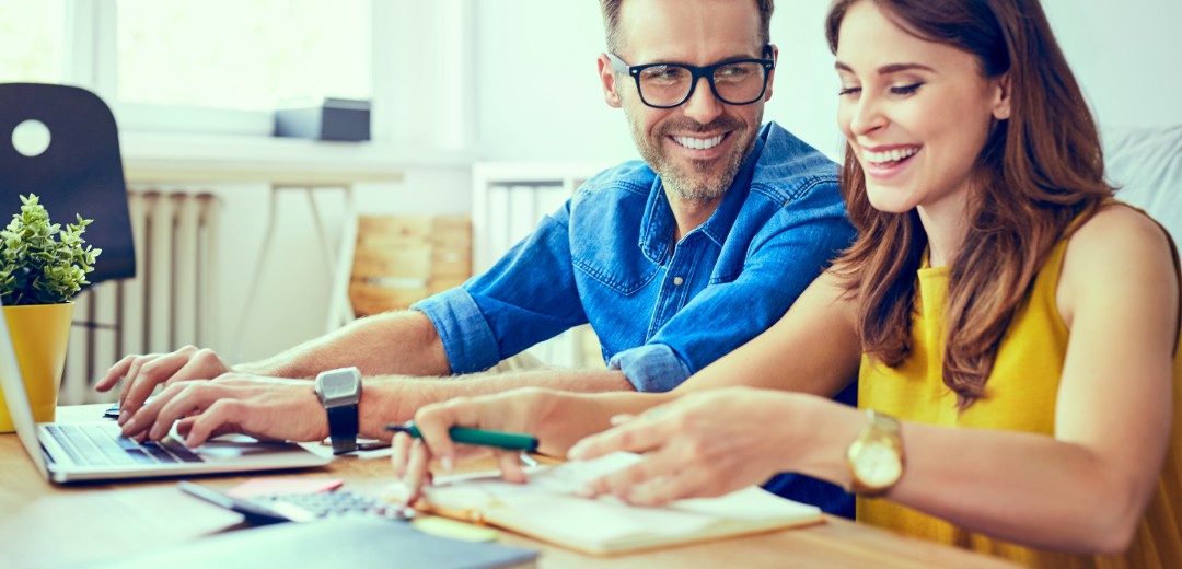 Ein Mann und eine Frau sitzen nebeneinander und unterhalten sich lachend, während sie an Unterlagen und am Laptop arbeiten. 