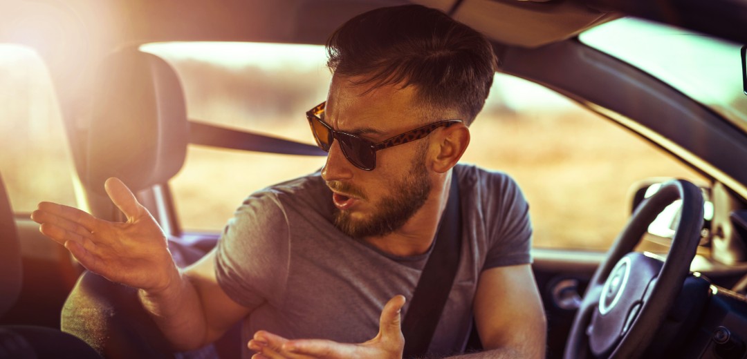 Ein Mann mit Sonnenbrille schaut verwundert und Schultern zuckend um sich im Auto.  