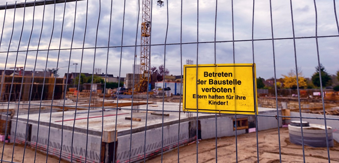 Das Bild zeigt eine Baustelle, welche mit einem Zaun abgesperrt ist. An dem Zaun hängt ein gelbes Warnschild, auf dem "Betreten der Baustelle verboten! Eltern haften für ihre Kinder!" steht. 