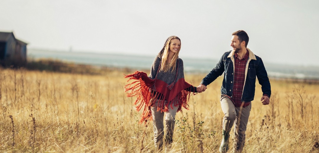 Eine junge Frau und ein junger Mann gehen lächelnd Hand in Hand über eine Wiese.
