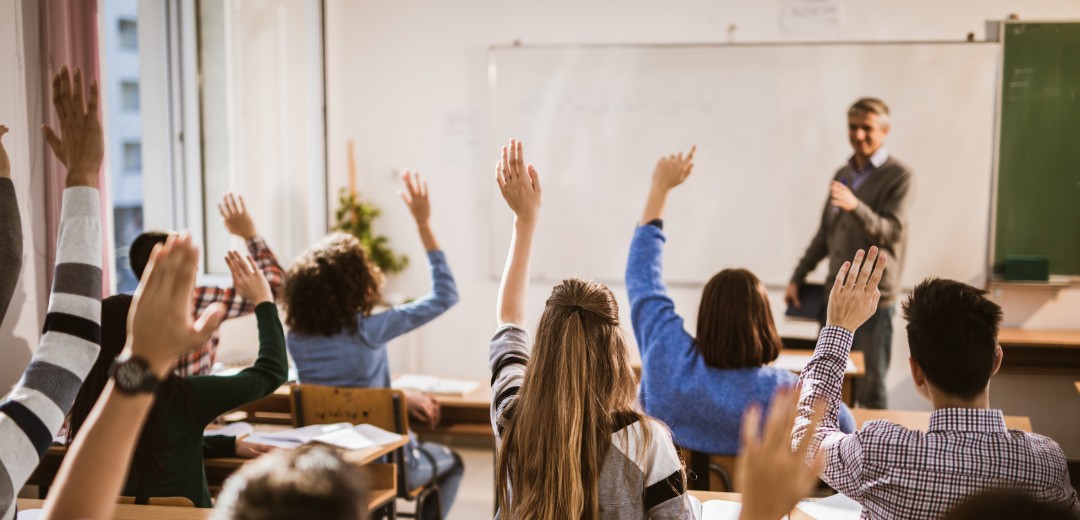 Ein Lehrer steht vor eine Klasse in einem Klassenzimmer. Viele Schüler melden sich. 