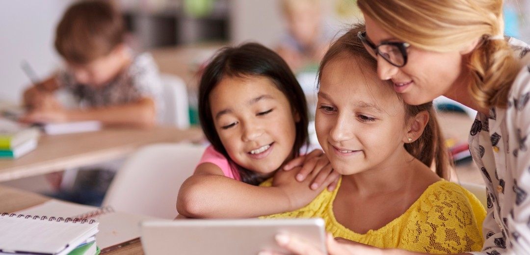 Eine Lehrerin zeigt zwei jungen Schülerinnen etwas auf dem Tablet. Sie sitzen in einem Klassenzimmer.