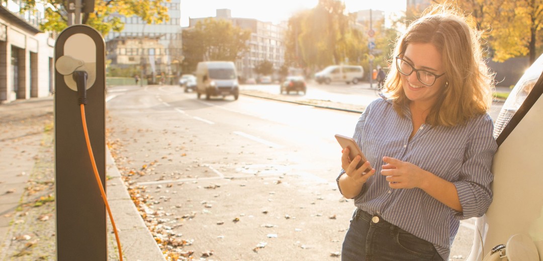 Eine Frau lehnt sich an ihr Auto, welches gerade auflädt, und schaut auf ihr Smartphone.