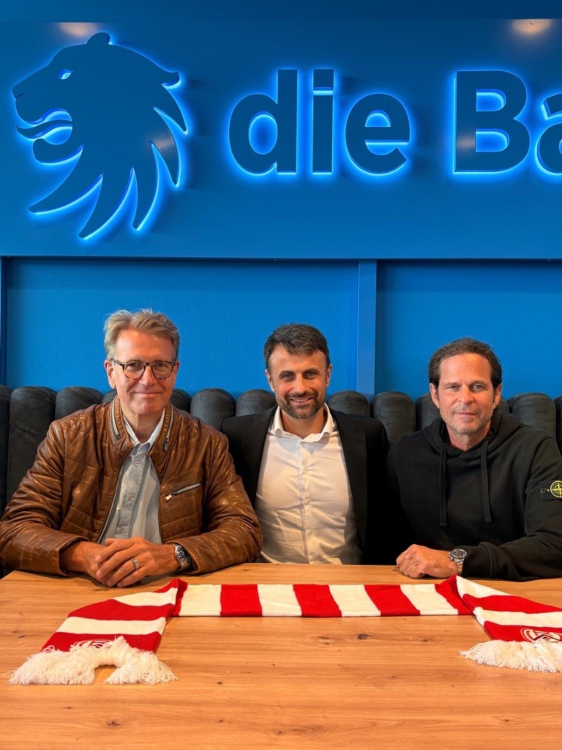 Drei Männer sitzen gemeinsam an einem Tisch vor dem beleuchteten Logo der Bayerischen; vor ihnen liegt ein rot-weißer Fanschal.